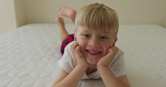 A blond boy lying on a bed in red pajama pants and a white T-shirt looks off to the side and smiles as he thinks about something.