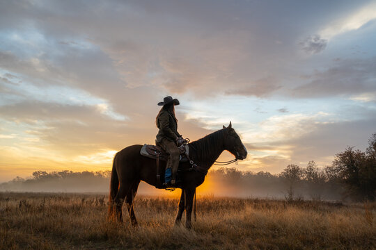 Woman Wearing Cowboy Hat Riding Horse and Looking Off At Golden Hour of Sunset With Cloudy Sky