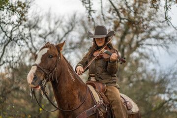 Fototapeta premium Cowgirl Woman Wearing Cowboy Hat Looking Down and Playing Fiddle Violin While Sitting On Brown and White Horse In Front of Trees and Cloudy Sky