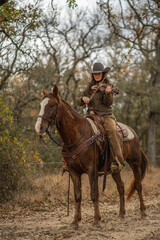 Fototapeta premium Cowgirl Woman Wearing Cowboy Hat Looking Down and Playing Fiddle Violin While Sitting On Brown and White Horse In Front of Trees and Cloudy Sky