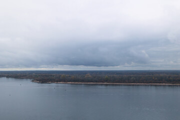 Obraz premium A Calm Landscape Of A Vast River Under A Heavy Overcast Sky. The Horizon Line Separates The Grey Water From The Autumn Forest Strip.