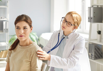 Serious female doctor in white medical uniform using stethoscope listening to patient heartbeat or lungs sounds back side, woman GP or physician consulting client meeting in modern hospital room