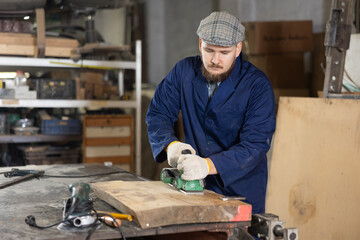 Young man worker in carpentry workshop polishes wooden board with machine