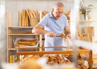 Senior man in bakery is picking up buns for tea. Client uses tongs to take croissants out of glass display case and put them in transparent disposable bag, collects set of sweet snacks for tea