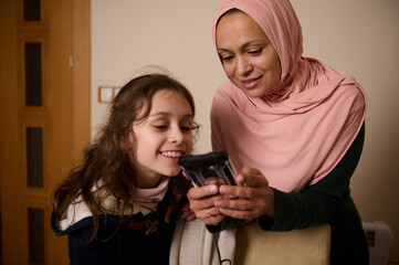 Mother And Daughter Share a Smile as a Muslim Woman in Hijab Looks at a Phone at Home