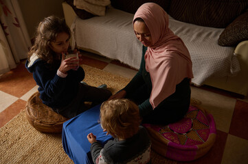 Mother In Hijab Plays With Kids At Home, Sharing Tea And Care In Cozy Living Room