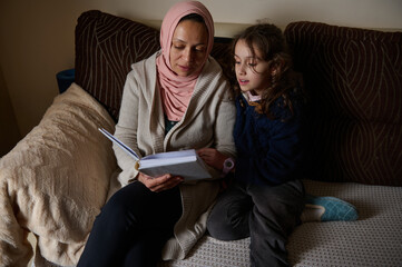 Mother in Hijab Reading With Daughter on Couch, Quiet Home Moment Together