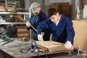 Carpenter measures the length of board with a tape measure. Foreman prompts him
