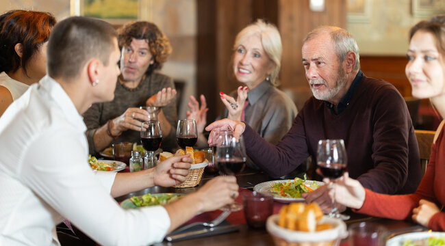 Large friendly family gathered at table to have lunch in restaurant. Relatives eat, drink wine, and socialize. People participate in discussion during meeting.
