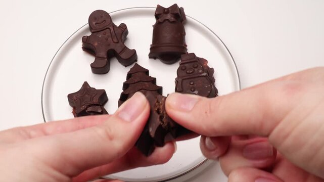 Hands breaking a holiday-themed chocolate candy with visible filling over a plate of assorted festive sweets shaped like gingerbread men, trees, and bells