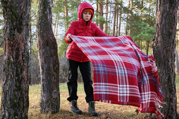 A young woman spreads a plaid blanket in the forest under a tree. Picnic in the park, outdoors in the forest