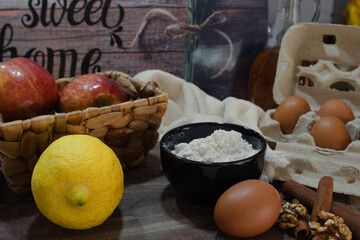 A variety of baking ingredients, including red apples, lemons and walnuts arranged on a rustic table, with a woven basket in the background. A traditional home cooking concept.
