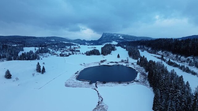 Aerial view of the dark waters of Lake Ter contrasting against the pristine white snow covering the landscape, Le Lieu, Vaud, Switzerland.