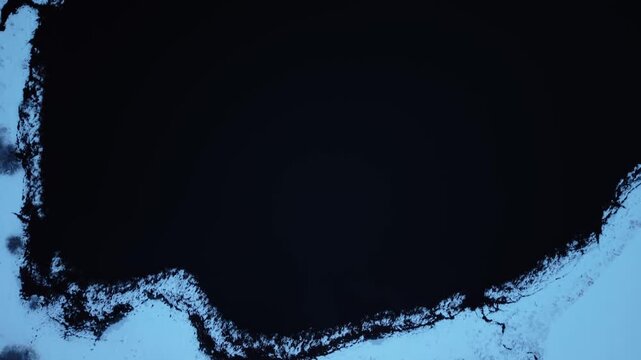 Aerial view of the dark, still waters of Lake Ter contrasting with the snow-covered shoreline, creating a serene, almost ethereal landscape, Le Lieu, Vaud, Switzerland.