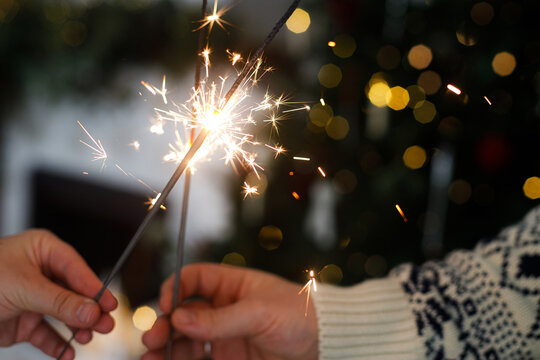 Hands holding fireworks against christmas tree and fireplace in festive decorated cozy living room. Happy New Year! Atmospheric family celebration with burning sparklers in hands close up