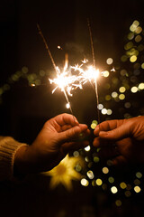Hands holding fireworks against christmas tree lights bokeh in evening cozy living room. Happy New...