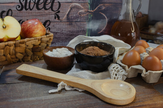 Tasty fresh ingredients including apples, cocoa powder, flour, and eggs arranged on a rustic wooden table, preparing for a homemade recipe or dessert creation
