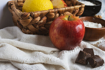 Baking ingredients including a red apple,lemons and dark chocolate arranged on a rustic linen tablecloth with a woven basket in the background. Traditional home cooking concept