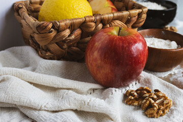 Fresh ingredients lay on rustic fabric with a red apple, walnuts, lemon, flour bowls, and a woven basket, preparing for a homemade dessert recipe