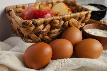 Fresh baking ingredients on a rustic table, whole brown eggs , bowls of flour and a basket of red apples ready for homemade pastries