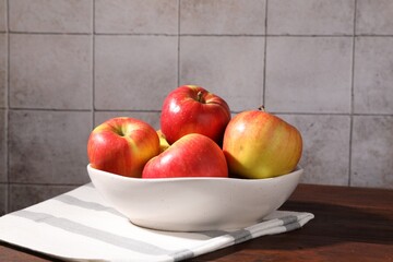 Ripe red apples in bowl on wooden table near grey tiled wall, closeup