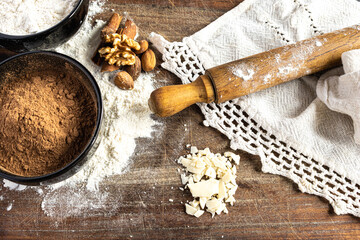 Fresh ingredients,cocoa powder, flour, mixed nuts and white chocolate on a wooden board with a rolling pin and linen cloth, ready for homemade festive dessert preparation
