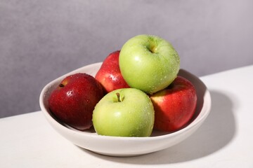 Fresh red and green apples with water drops in bowl on white table against grey background, closeup