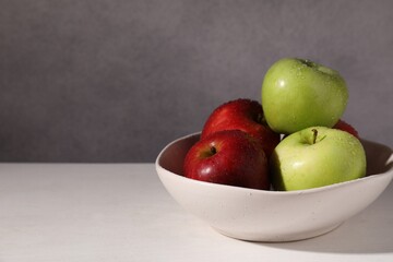 Fresh red and green apples with water drops in bowl on white table against grey background, closeup. Space for text