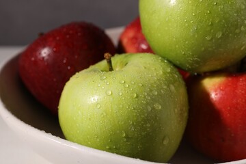 Fresh red and green apples with water drops in bowl on white table against grey background, closeup