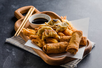 Asian appetizers including a crispy egg roll, fried cream cheese wontons, and a pork dumpling (potsticker) served with soy sauce on a dark textured background.