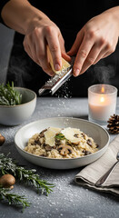 Creamy mushroom risotto with parmesan flakes, hands grating fresh parmesan cheese directly over the bowl, surrounded by snow-dusted rosemary, ceramic bowl, frosty candle glass and napkin