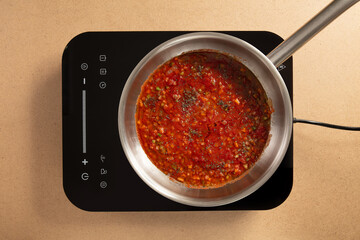 A pot of fresh tomato sauce simmers on an induction cooktop, blending diced tomatoes with herbs and spices. The modern kitchen setting highlights the sleek technology.