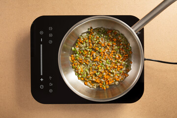 Fresh diced vegetables, including carrots, onions, and peppers, are sautéing in a stainless steel pan on a sleek induction cooktop. The kitchen offers a clean and inviting atmosphere.
