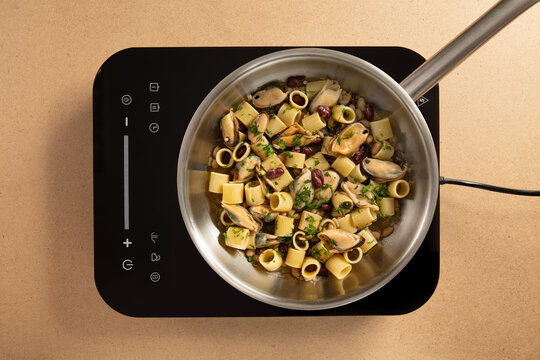 A pot with pasta, mussels, and beans simmers on an induction cooktop in a modern kitchen. Fresh herbs are sprinkled on top, adding color and flavor to the meal.