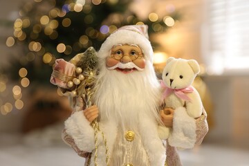 Santa Claus figure with sack of gifts and toy bear against blurred lights indoors, closeup. Merry...