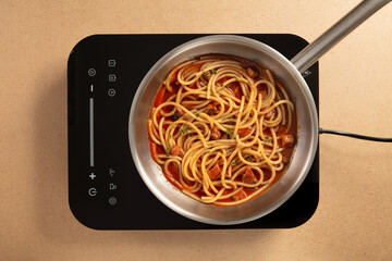 Long strands of spaghetti simmer in a rich tomato sauce in a stainless steel pot on an electric cooktop. The warm kitchen space invites a sense of comfort and home cooking.