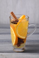 Delicious mulled white wine with slices of orange and spices in glass cup on grey wooden table, closeup