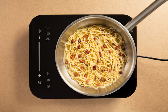 A pot of spaghetti is seen cooking on a stylish induction cooktop, featuring crispy bacon pieces mixed in a rich, creamy sauce, in a modern kitchen environment.