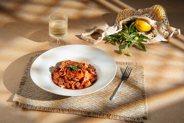 Pappardelle pasta in rich tomato sauce with meat and basil on a rustic placemat. Warm sunlight, wine and lemons add a cozy Mediterranean home-cooking atmosphere.