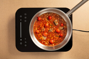 A pot filled with a rich stew simmers on an induction cooktop, showcasing vibrant tomatoes and tender meat. Fresh herbs and spices add flavor to the dish.