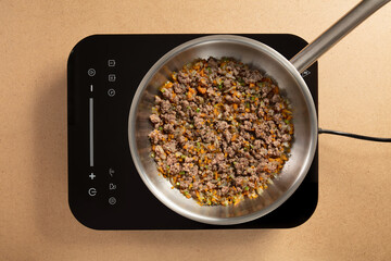 Ground meat is being cooked with a mix of chopped vegetables on a sleek electric stovetop. The scene shows a contemporary kitchen environment, highlighting freshness and preparation.