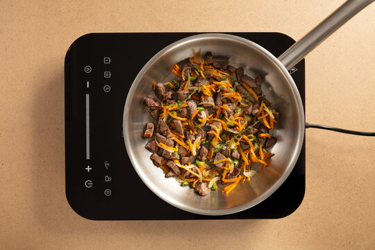 Beef and colorful vegetables are being sautéed in a stainless steel pan on a sleek induction stove. The kitchen has a clean, contemporary design with good lighting.