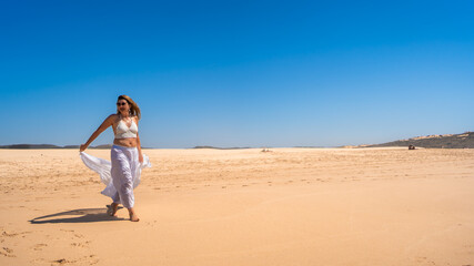 Beautiful middle-aged woman walking holding scarf blowing in wind on sandy beach Bordeira Portugal in summer. Front view	