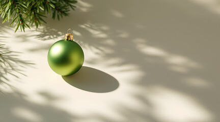 Minimalistic holiday scene with a single green Christmas ornament casting a soft shadow on a light surface. Natural sunlight and evergreen branches create a clean, elegant seasonal mood perfect for mo