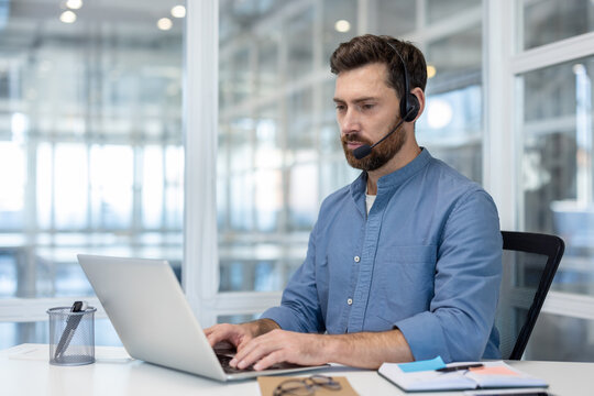 Bearded male agent working at laptop, wearing headset with microphone, providing customer service and technical support in a modern office environment