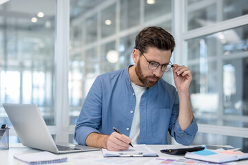 Serious male professional analyzing financial data, calculating expenses, and taking notes on...