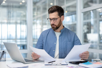 Young businessman wearing glasses carefully reviewing financial paperwork and working with laptop at desk, managing important financial data and administrative tasks in an office environment
