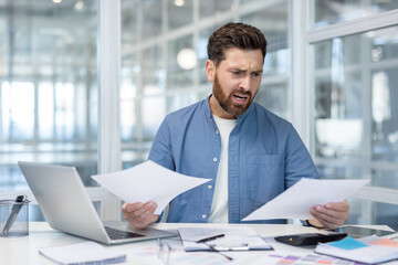 Man frustrated and stressed while reviewing bills and paperwork at a modern home office desk, worried about finances, debt, taxes and payments on laptop and calculator
