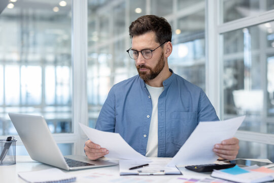 Young businessman wearing glasses carefully reviewing financial paperwork and working with laptop at desk, managing important financial data and administrative tasks in an office environment