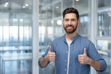 Bearded man smiling broadly and wearing a blue shirt extends both thumbs up, expressing approval,...